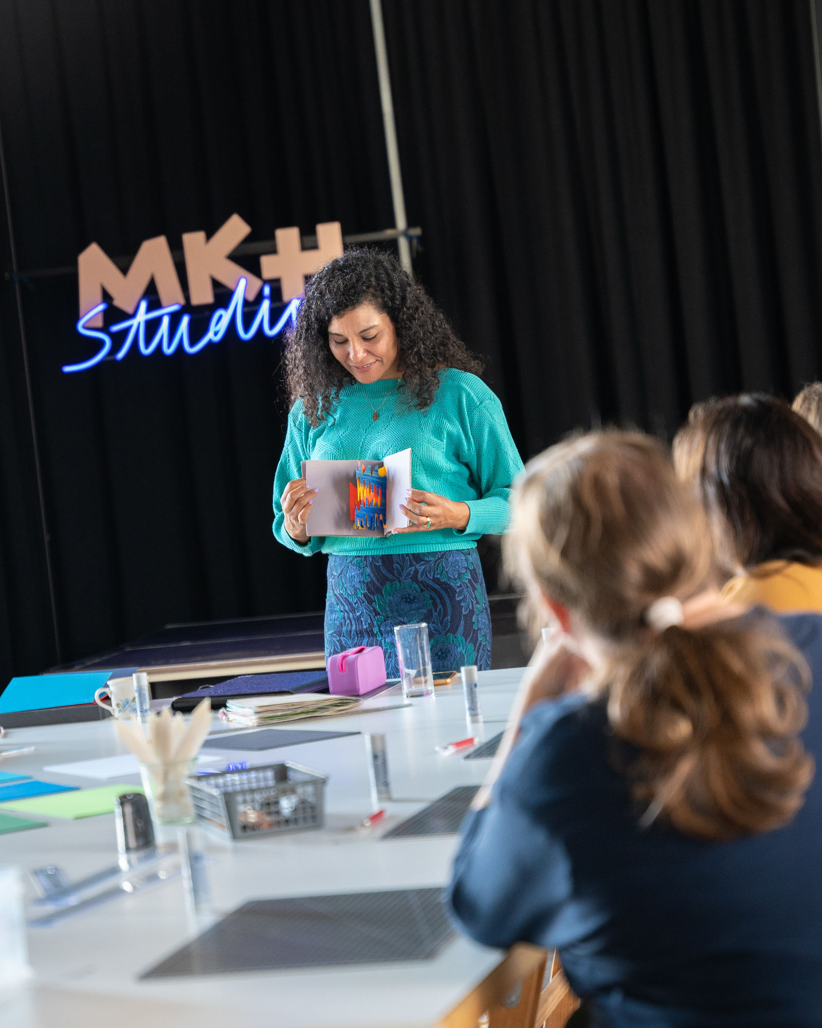 Frau steht neben Tisch und hält ein Papier-Pop-up Buch in beiden Händen. Am Tisch Liegen Papier- und Schneideutensilien. Leute sitzen am Tisch, den Blick zu ihr gerichtet.