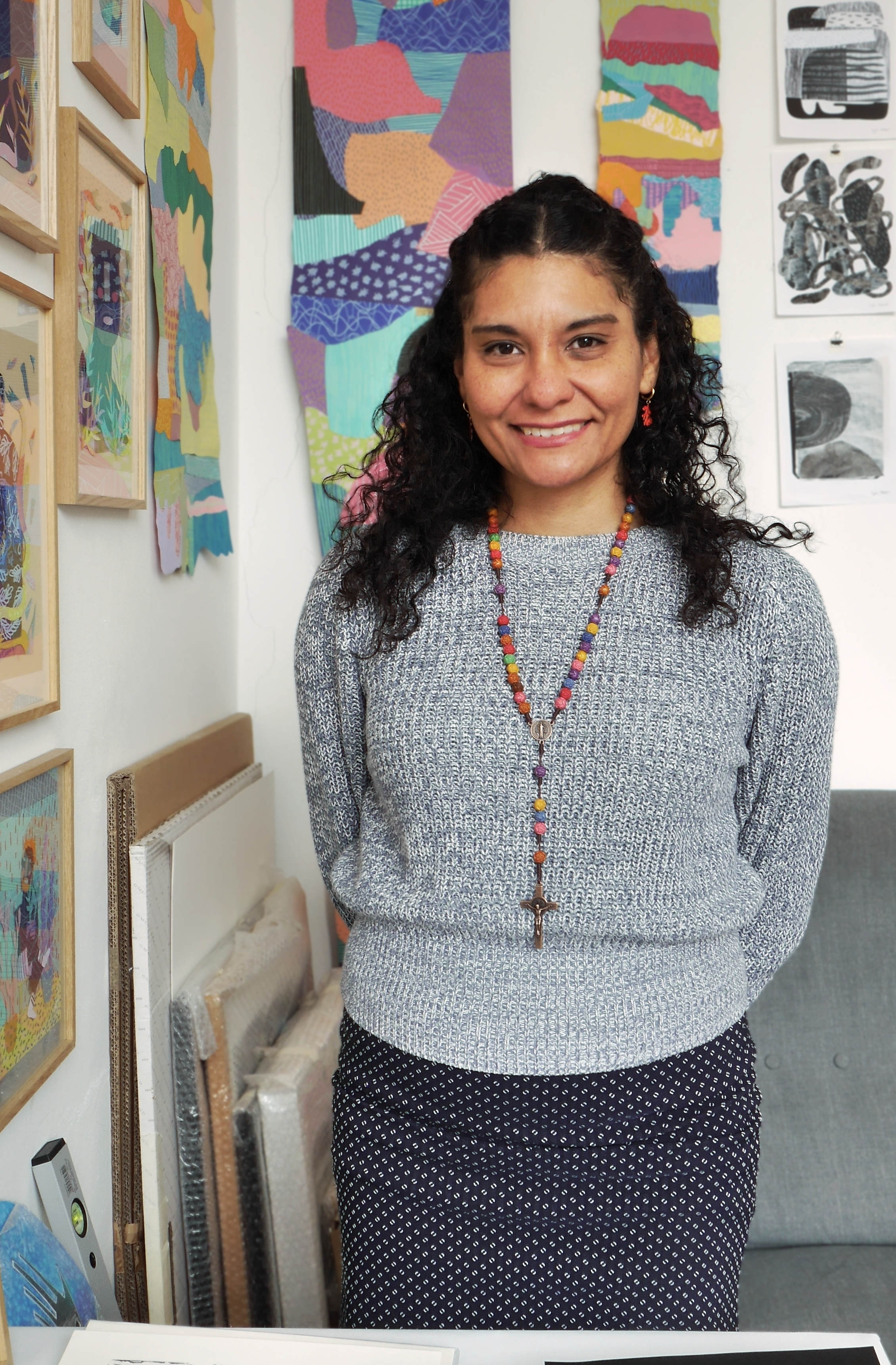 Frau steht lächelnd in einem Atelier. An Wand hängen verschiedenste Kunstwerke aus Papier.